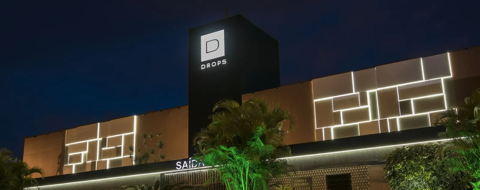 Fachada de uma unidade da Rede Drops à noite: o totem com o logotipo iluminado ao centro e a parede de luz ao lado