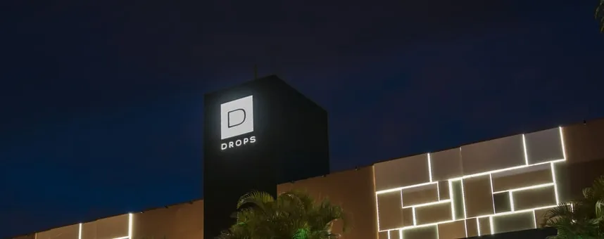Fachada de uma unidade da Rede Drops à noite, com o letreiro iluminado e a parede de luz